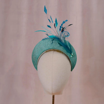A teal  hat in a headband style with tonal teal feather and bead accents displayed on a teal mannequin  against a soft pink backdrop. Ideal for weddings, royal garden parties and horse racing. 