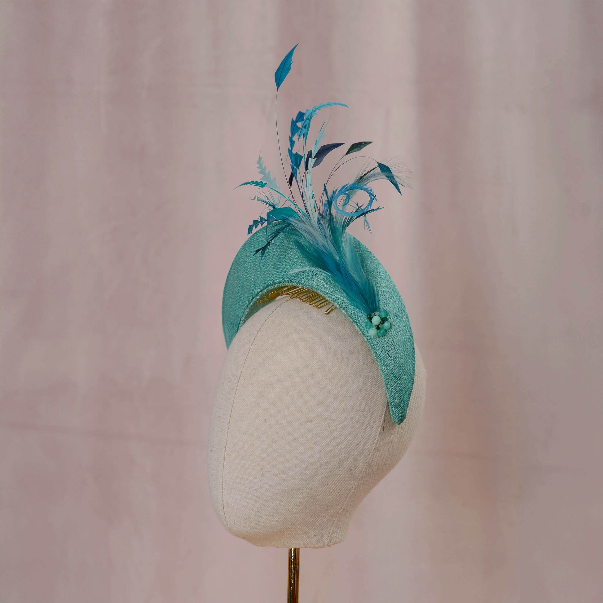 A teal and turquoise feather fascinator headpiece displayed on a cream fabric mannequin head against a soft pink backdrop.