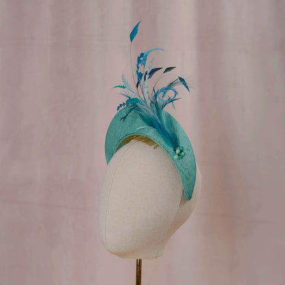A teal and turquoise feather fascinator headpiece displayed on a cream fabric mannequin head against a soft pink backdrop.