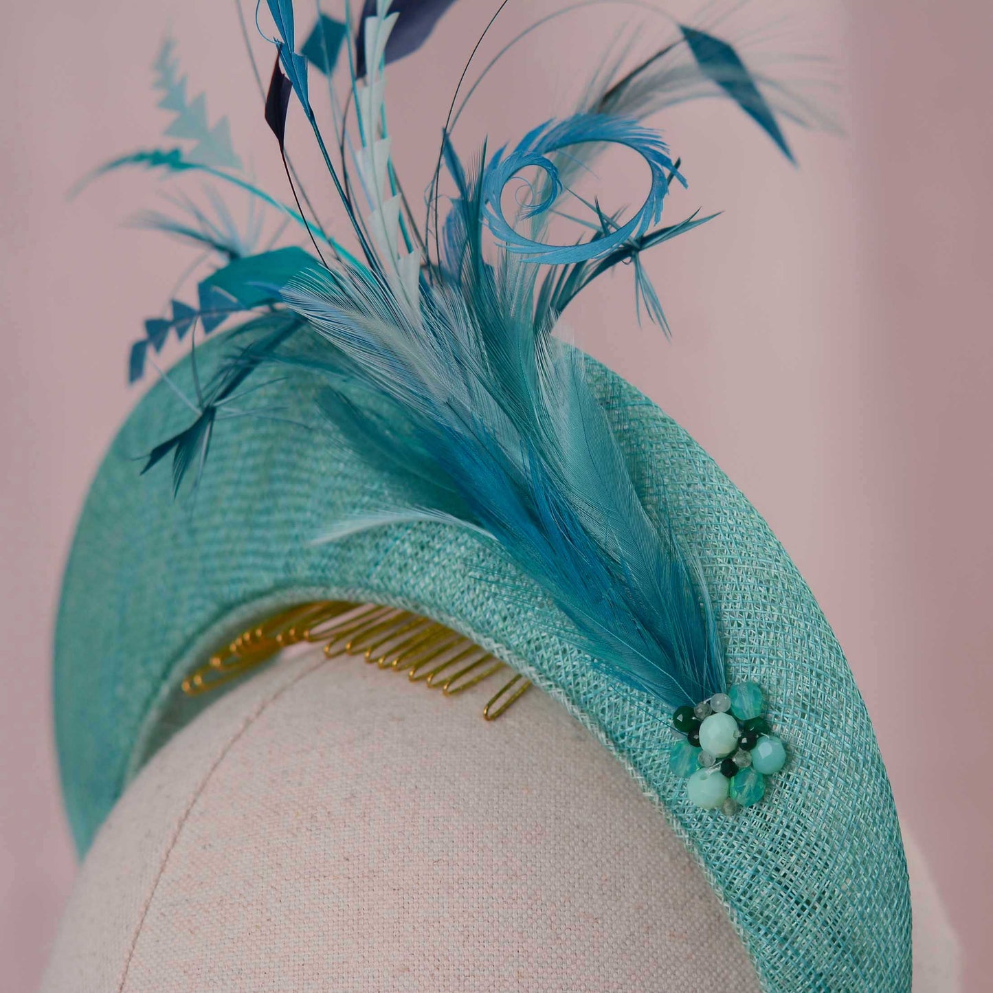 A close-up of a textured teal and turquoise feather fascinator headpiece with a gold comb and a small bead cluster on a mannequin display.