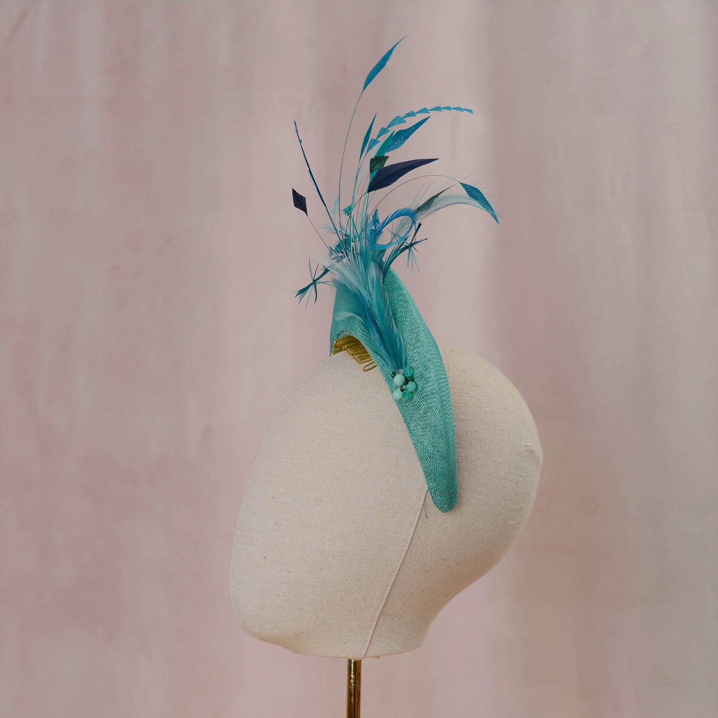 A turquoise and teal feather fascinator headpiece displayed on a cream fabric mannequin head against a soft pink backdrop.