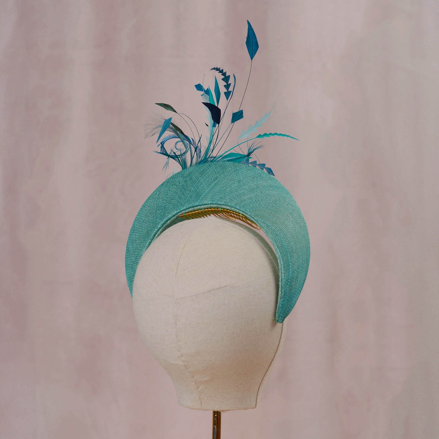 A turquoise blue fascinator hat with decorative feathers and a gold comb, displayed on a white fabric mannequin head against a soft pink background.