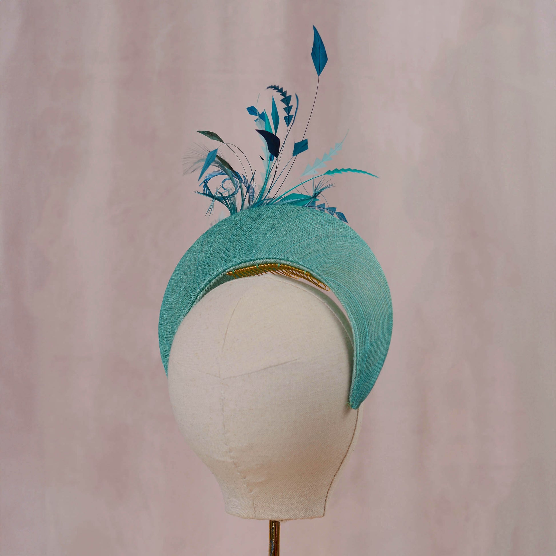 A turquoise blue fascinator hat with decorative feathers and a gold comb, displayed on a white fabric mannequin head against a soft pink background.