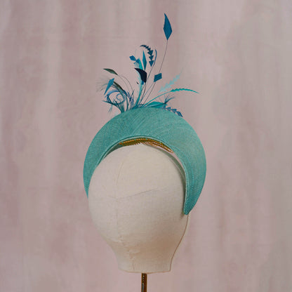 A turquoise blue fascinator hat with decorative feathers and a gold comb, displayed on a white fabric mannequin head against a soft pink background.