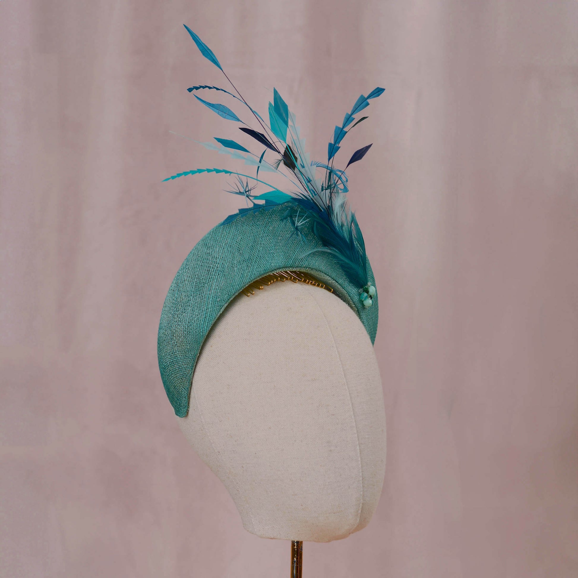 A teal fascinator hat with matching feathers and bead accents, displayed on a cream mannequin head against a soft pink backdrop.