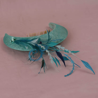 A curved mint green woven straw fascinator headband with a  gold-tone metal comb set, decorated with a teal blue feather sand a small cluster of light green and turquoise beads, set against a dusty pink background.