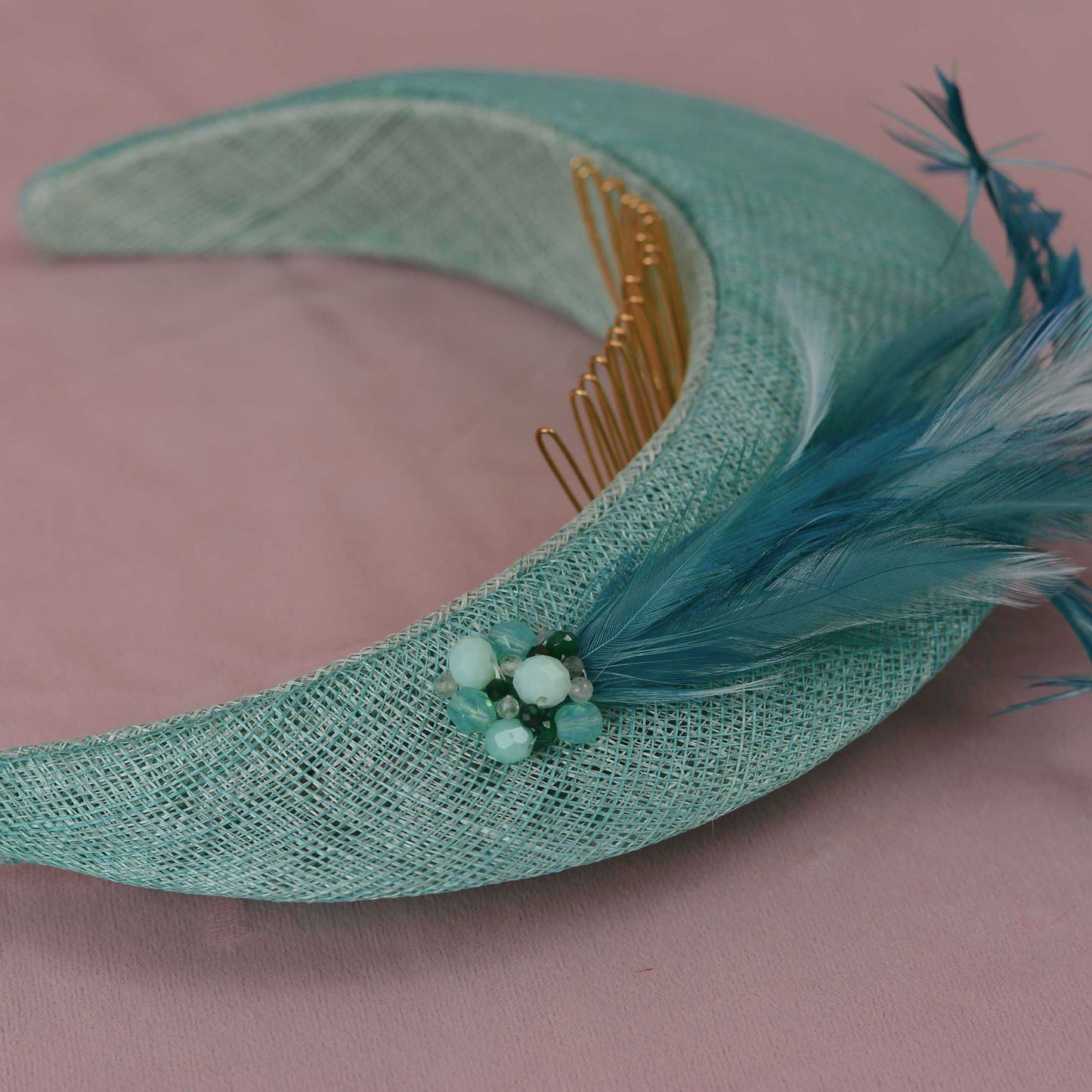 A curved mint green woven straw fascinator headband with a  gold-tone metal comb set, decorated with a teal blue feather sand a small cluster of light green and turquoise beads, set against a dusty pink background.