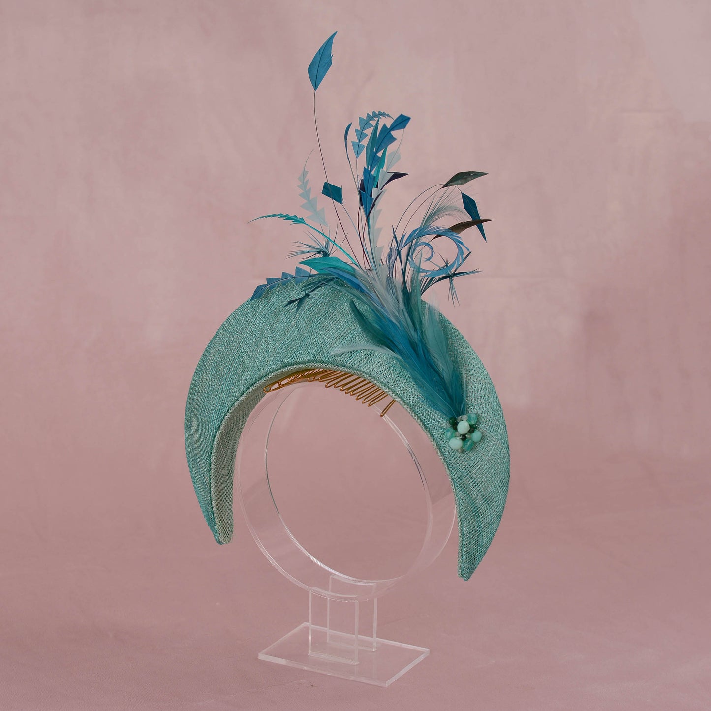 A structured fascinator hat in textured turquoise green with a large feathered spray in shades of blue, teal, and green, with matching beading displayed on a clear acrylic stand against a dusty pink background.